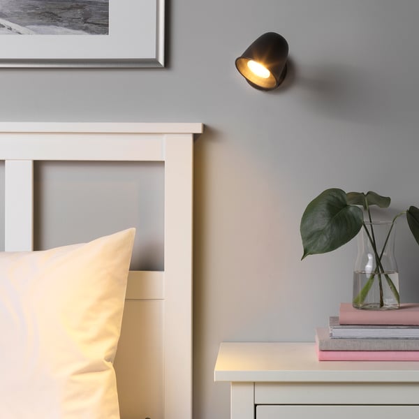 Small, black, adjustable wall light, SKURUP, highlighting a cosy room corner with a bed and books.