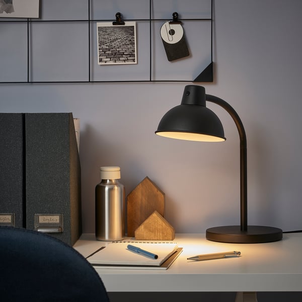 Minimalist desk setup with a small black SKURUP lamp, emit bright light, surrounded by books and decor.