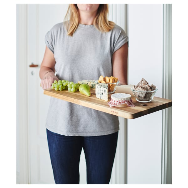 Person holds wooden serving tray with fruits, cheeses, and crackers in a light-colored room.
