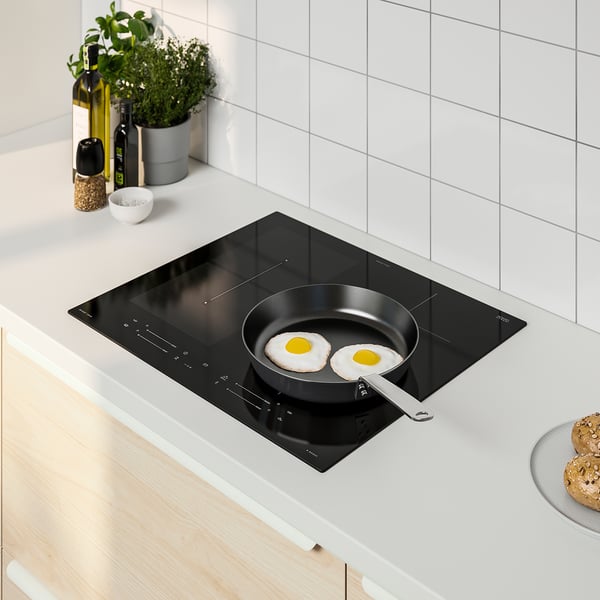 Modern kitchen with SMAKLIG black induction cooktop, sunny-side-up eggs frying, sleek design, wooden spice rack, plant, cookies, cosy vibe.