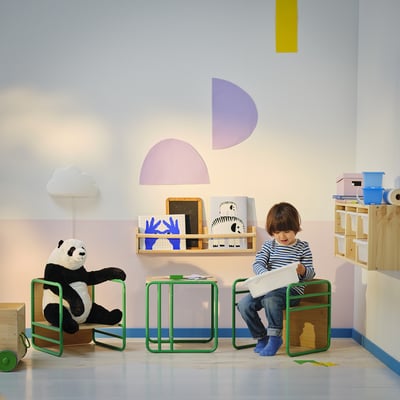 Kids playroom with SMÅSKRAKE chairs, one green, one with stuffed panda, boy sits on green chair, pastel wall art, books, and toys.