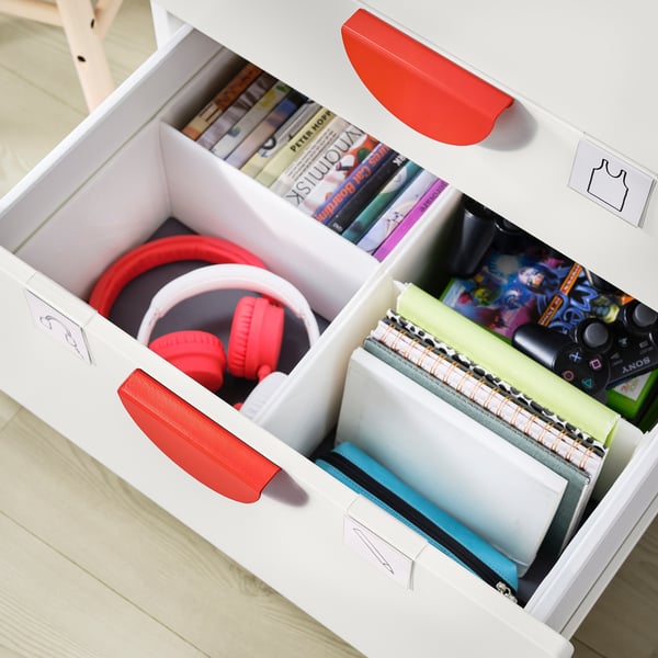Open white storage unit showing organised drawer with red headphones, green and grey notebooks, and colourful game cases.