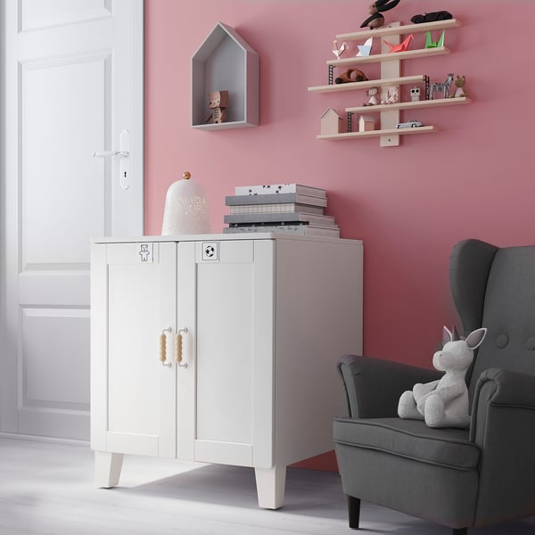 A childrens room with a white SMÅSTAD storage cabinet, grey chair, books, toys, and wall shelves against pink walls.
