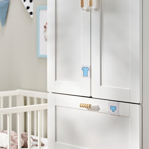 A white SMÅSTAD wardrobe with blue icons and wooden knobs, next to a white crib.