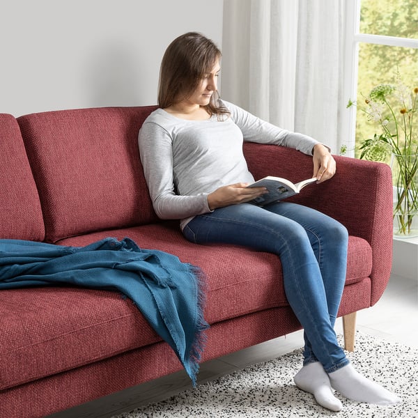 Woman reads book on red SMEDSTORP sofa with soft, retro style and modern comfort, near window.