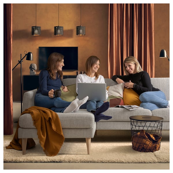 Three people relax on a sofa in a cosy living room. One works on a laptop, another drinks coffee, and the third reads.