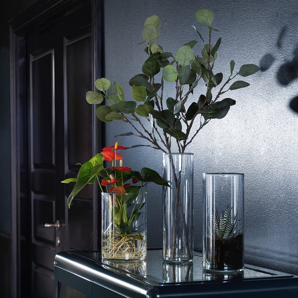 Three clear vases on a counter, two with green leaves, one with red flowers, all modern and minimalist, part of the SMYCKA collection.
