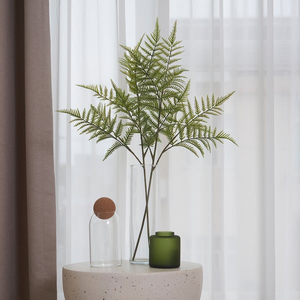 A clear vase holds a fake fern on a speckled table, with a candle nearby.