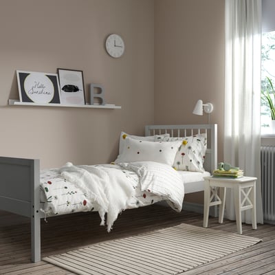 Childs bedroom with grey SMYGA bed, floral sheets, bedside table, clock shelf, and sheer curtains.