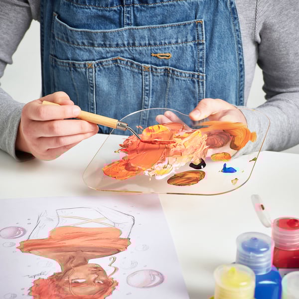 Mixing paint on palette with spatula, art supplies on table, canvas nearby.