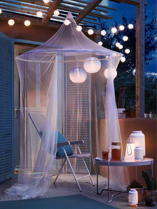 Relaxing patio scene with a white SOLIG net canopy, string lights, blue chairs, and table holding drinks and lanterns.