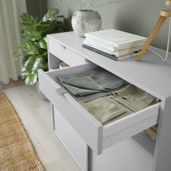 White cabinet with open drawer, folded clothes inside, on top, books, plant, lamp, green wall.