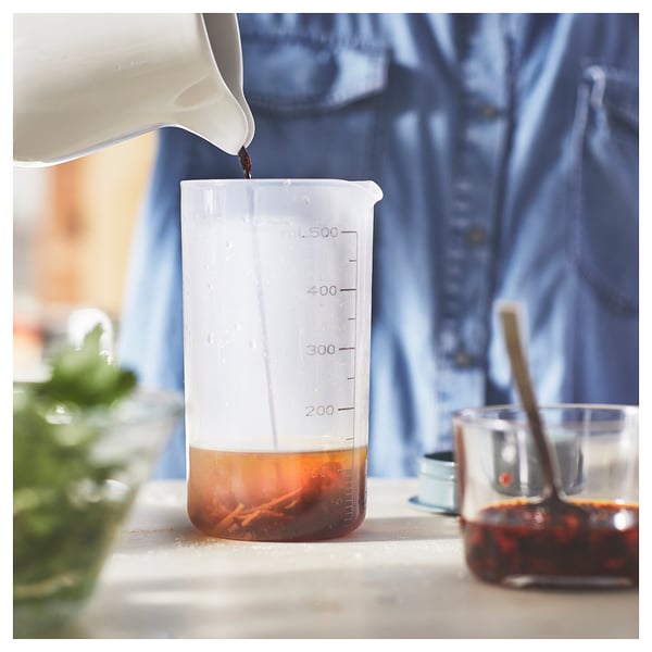 Measuring cup fills with dark liquid from pitcher, nearby bottle and herbs.