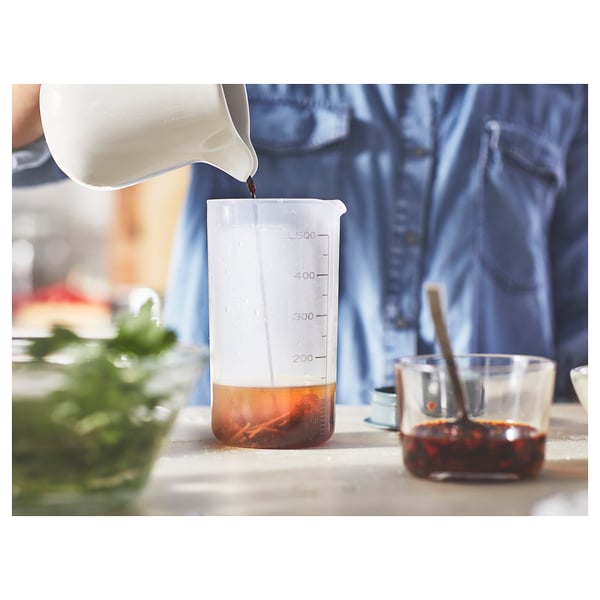 Person pouring liquid into shaker with markings, next to spoon in red liquid, indicating food prep.