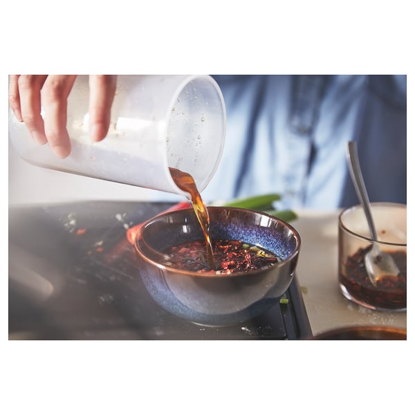 Hand pours liquid from a clear plastic pitcher into a blue glass bowl on a stove. Liquid is amber colored.