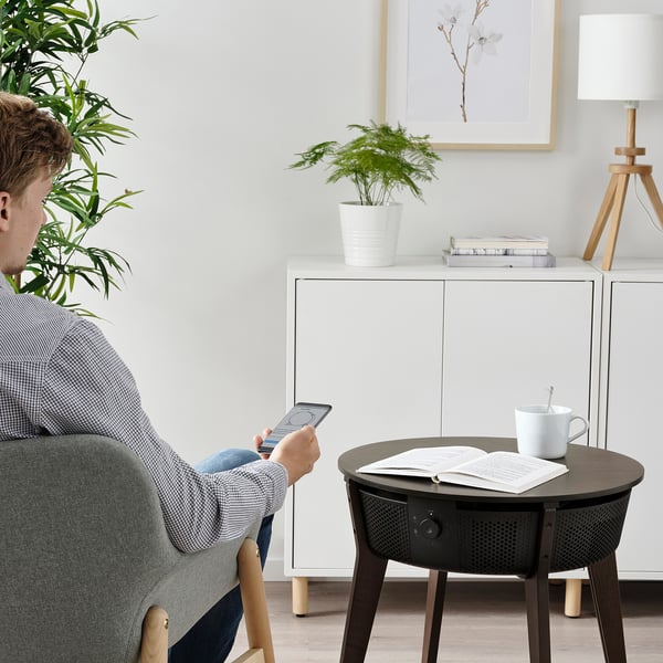 A person sits in a chair, using a smartphone. Nearby, a STARKVIND air purifier table holds a book, cup, and remote, improving indoor air quality.