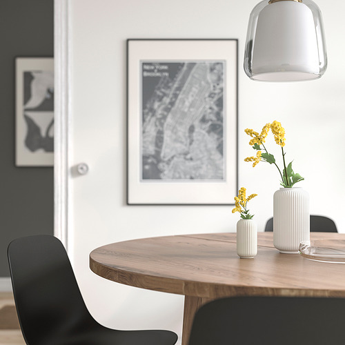 Small white STILREN vase with yellow flowers on wooden table near black chair, framed city map, and hanging light.