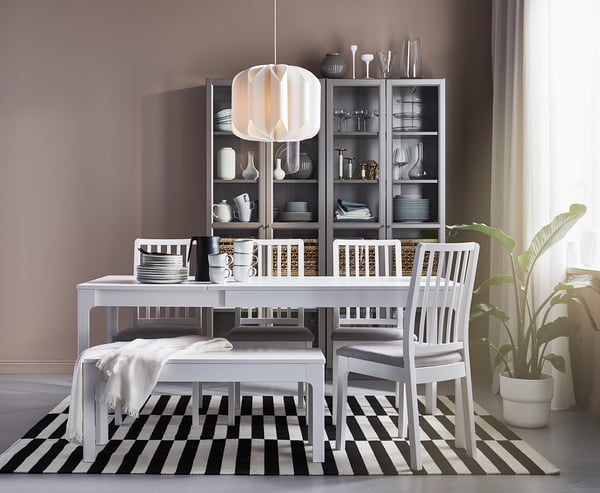 Modern dining area with white table, grey chairs, black-and-white striped rug, grey walls, shelves with white dishes, hanging lamp, and potted plant.