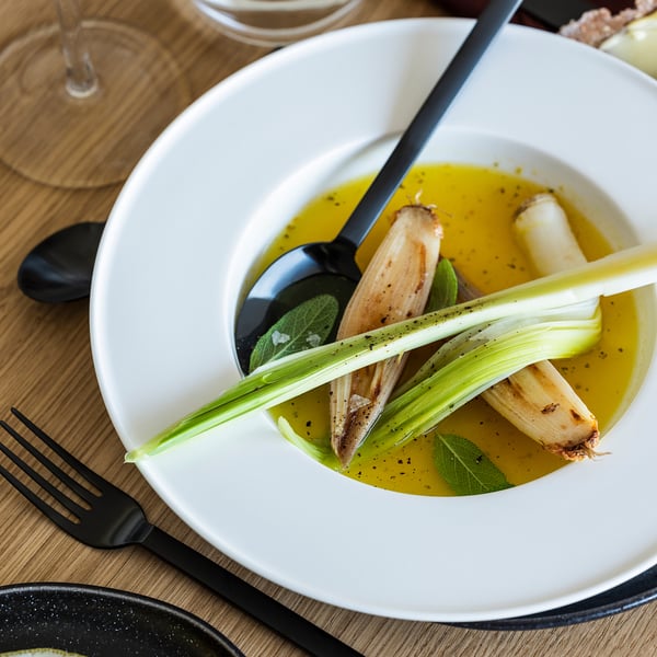 White deep plate with roasted vegetables and sauce, accompanied by black cutlery.