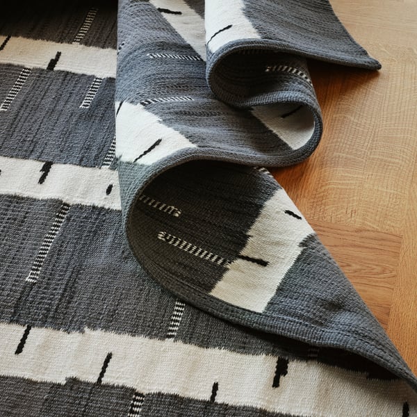 Black and white handwoven wool rug with birch tree pattern partially rolled on wooden floor. High-quality craftsmanship visible in detailed texture.