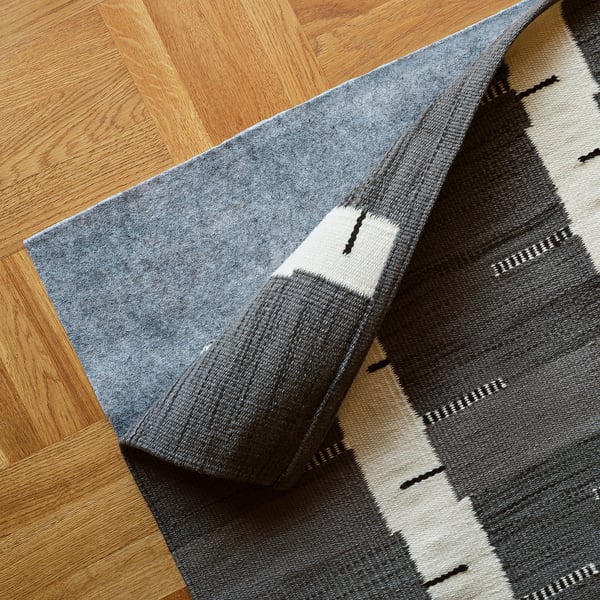 A grey handwoven rug with a birch tree pattern lies on a wooden floor. It appears reversible and flat-woven for easy use under furniture.