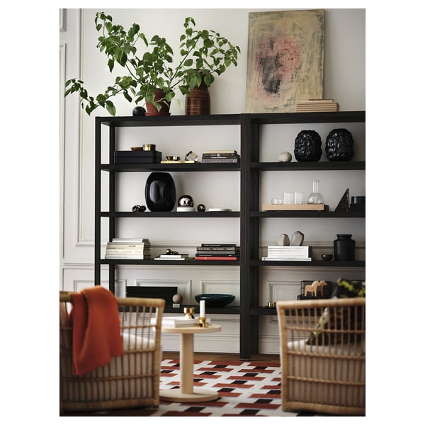 Stylish black STOCKHOLM 2025 shelves hold books & decor, larger than wicker chairs, with green plant atop.