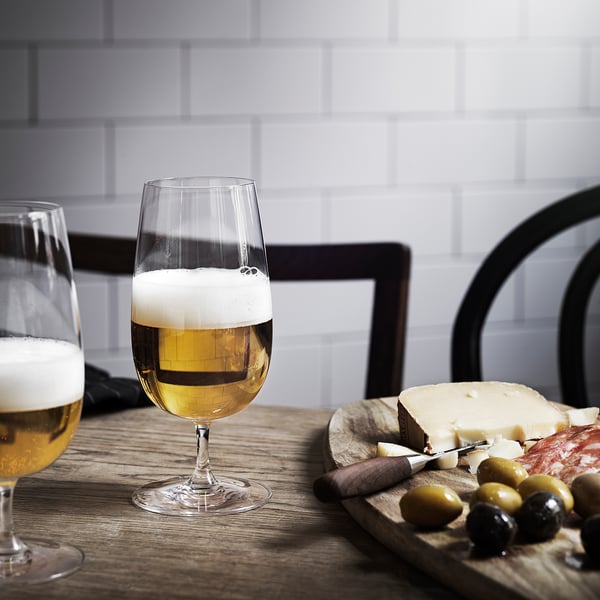 Two glasses of beer on a wooden table with a cheese and olive platter, set against a tiled wall with black chairs.
