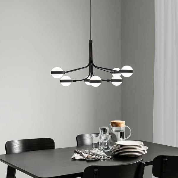 Modern black chandelier with 8 lights over a dark dining table with place settings and chairs against minimalist grey walls with a white curtain.