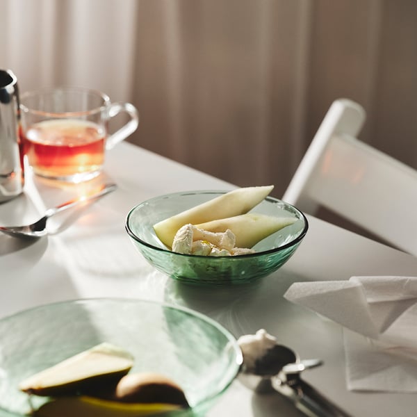 Bowl of sliced melon on a table, accompanied by a glass of tea, showcasing the STRANDKRABBA bowls inviting shape and practical stacking feature.