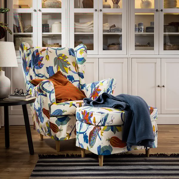 STRANDMON wing chair with floral pattern, blue throw, side table with lamp & books, white cabinet with dishes.