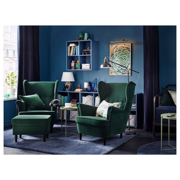 Green velvet wingback armchair with ottoman, dark blue walls, area rug, floor lamp, and shelves with decor.
