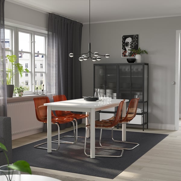 Modern dining room with STRANDTORP table, orange chairs, set for four, black sideboard, rug, large window, chandelier, painting, and plants.
