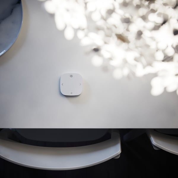 White circular remote control, centred on a light grey tabletop. Near a plant; light lorenzo.