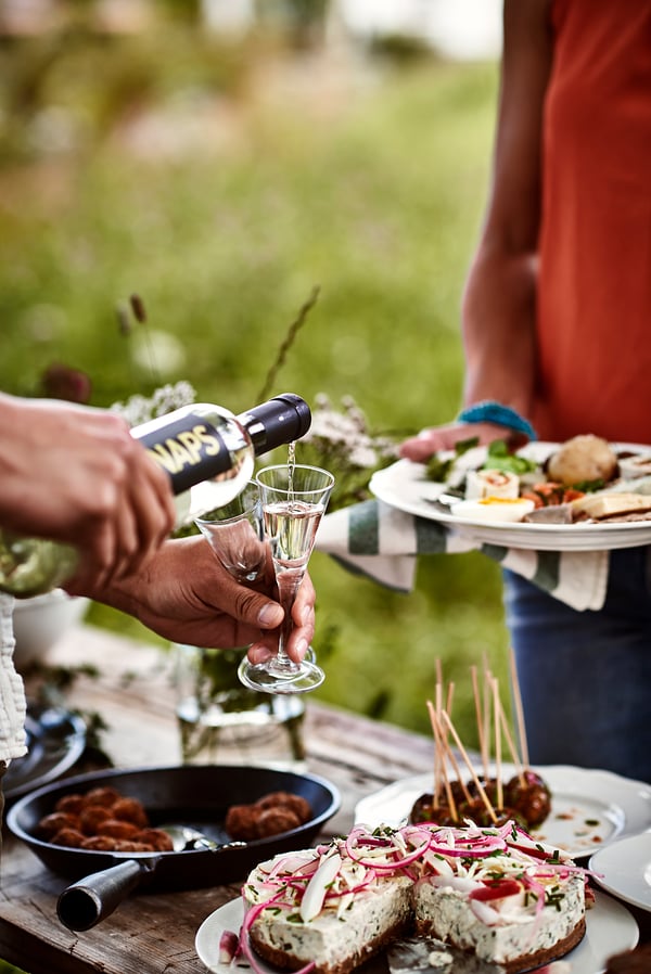 Person pouring drink from SVALKA bottle into wine glass. Outdoor meal with food laid out. Person in orange shirt holding plate.