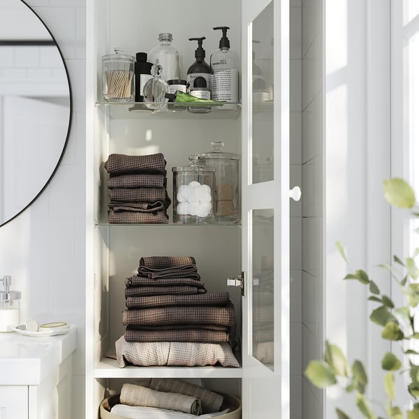 Tannforsen white glass-door cabinet stores folded towels and toiletries neatly on adjustable shelves, mounted high to save floor space.