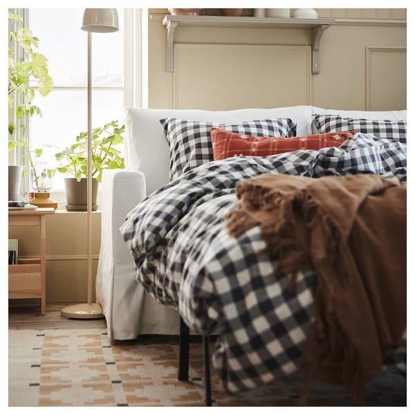 Bedroom with black and white chequered bedspread, large window with white blinds, floor lamp, plant, and a rug with a geometric pattern.