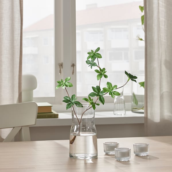 Small glass vase holds flowers on table by window. Two smaller glass cups sit beside it. Bright light fills the room through sheer curtains.