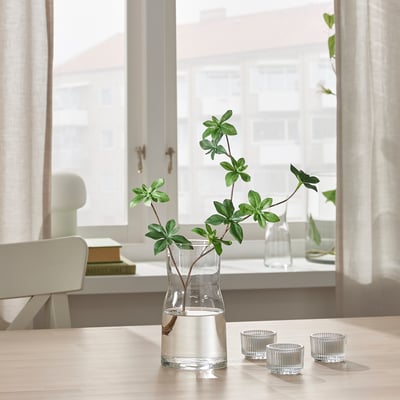 Small glass vase holds flowers on table by window. Two smaller glass cups sit beside it. Bright light fills the room through sheer curtains.