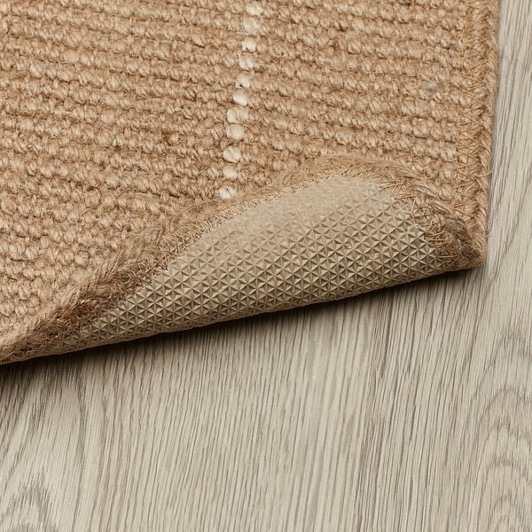 Corner of beige jute rug on wooden floor, showing textured non-slip backing.