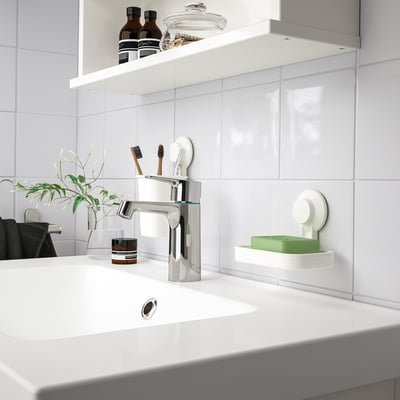 Modern white bathroom sink with chrome tap. Small TISKEN green soap dish, toothbrush holder, and plant. Clean, minimalist design.