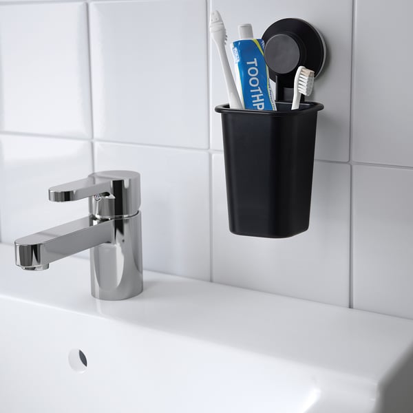Black TISKEN toothbrush holder with suction cups, holding toothbrushes and toothpaste near a chrome tap on a white sink, in a minimalist bathroom.