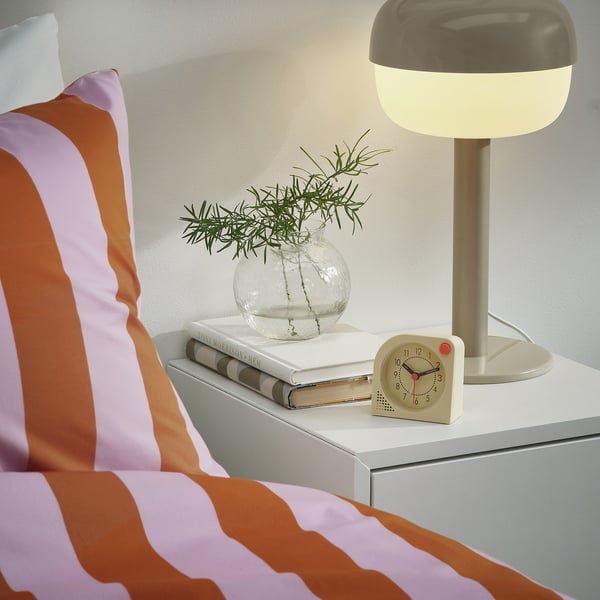 Striped bed with a bedside table. Table has books, a round vase with greenery, a clock with a light-up alarm, and a lit lamp.