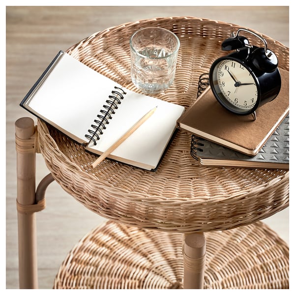 A TOLKNING natural rattan side table with a removable tray. The table holds a notebook, alarm clock, and glass.