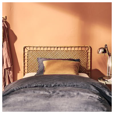 Natural rattan headboard with woven design, beige pillows, peach wall, grey bedding, lamp, coat hanger.