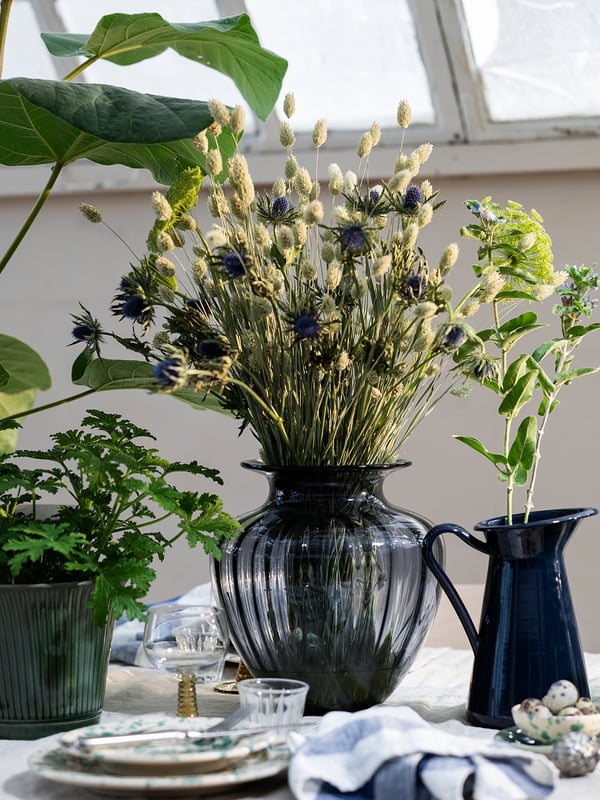 Glass vase with dried lavender and blue flowers, beside a blue pitcher on table with green plants and plates.
