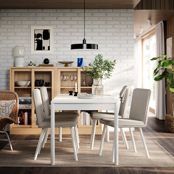 A TONSTAD dining set with off-white table, wooden legs, four chairs, bookshelf, vase, candles, and artwork against a brick wall.