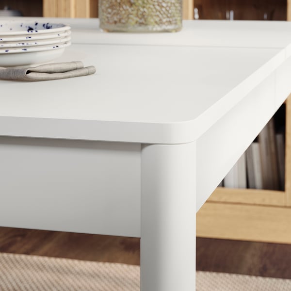 Simple, modern dining table. Clean, smooth white top with rounded edges. Sturdy legs, neat finish. Plates, napkins displayed.
