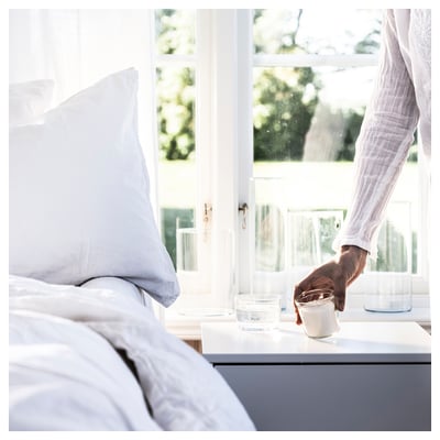 A person holds a glass near a window beside a white pillow. Sunlight streams in, illuminating the scene.