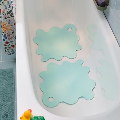 Two light turquoise TRAMPSJÖN bath mats with suction cups in a white tub beside a colourful shower curtain.
