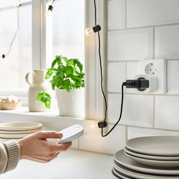 A hand holds a white remote near stacked plates, controlling a plug with a black cord. The scene includes a plant, plates, and a white wall.